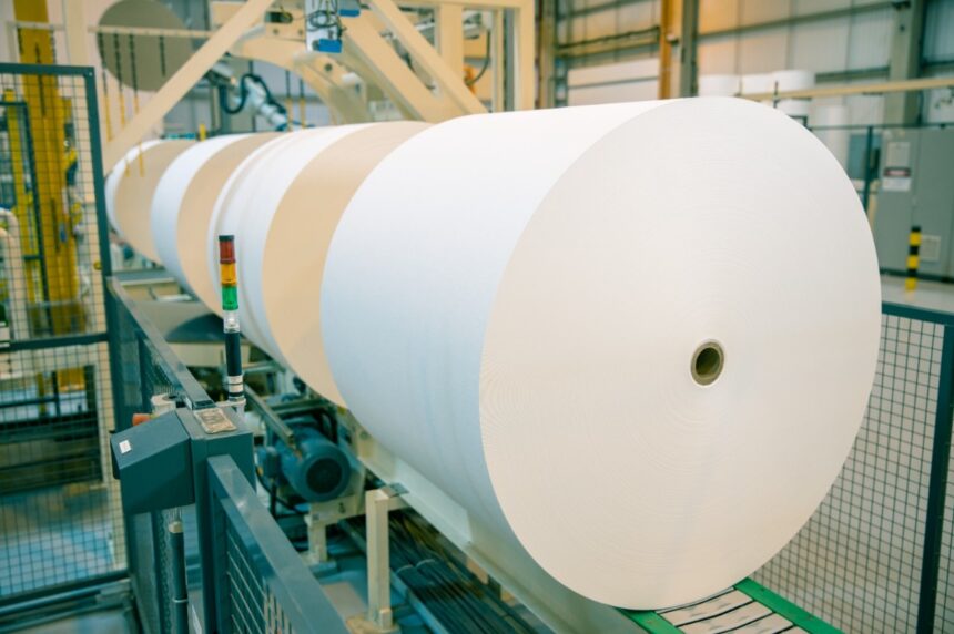 Grande rolo de papel branco em máquina industrial em fábrica, destacando processo de produção de papel e automatização.