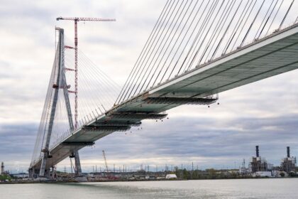 Ponte estaiada em construção sobre o rio, com guindaste ao fundo e céu nublado, destacando engenharia civil moderna.