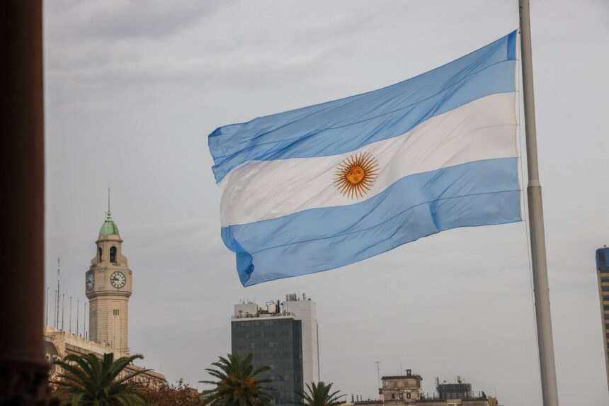 Bandeira da Argentina tremulando em frente a prédios históricos e palmeiras, simbolizando o patriotismo no centro urbano.