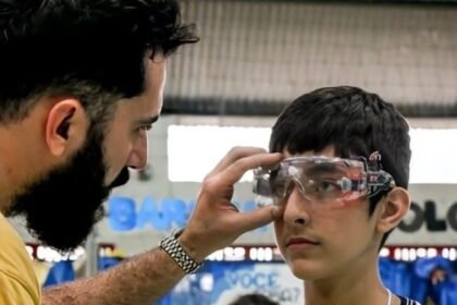 Homem ajusta óculos de proteção em jovem estudante em sala de aula, promovendo segurança no aprendizado prático de ciências.