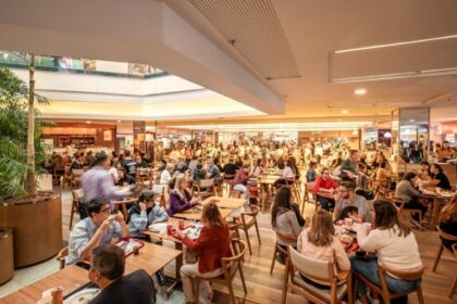 Praça de alimentação movimentada em shopping, com pessoas desfrutando refeições em mesas de madeira sob iluminação natural.