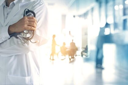 Médico com estetoscópio nos braços em corredor de hospital, mostrando atendimento e cuidados de saúde.
