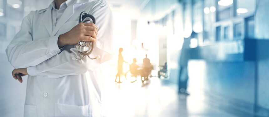Médico com estetoscópio nos braços em corredor de hospital, mostrando atendimento e cuidados de saúde.