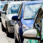 Carros estacionados em fila à beira de calçada em uma rua movimentada, refletindo o céu em seus para-brisas.