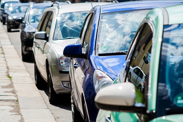 Carros estacionados em fila à beira de calçada em uma rua movimentada, refletindo o céu em seus para-brisas.