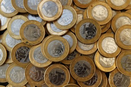 Várias moedas de um real, moeda brasileira, empilhadas, destacando o relevo metálico prateado e dourado.