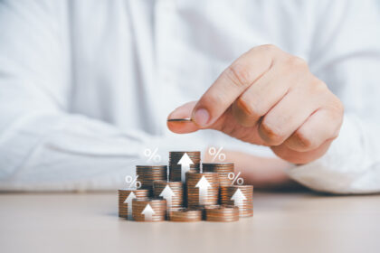 Mão coloca moeda em pilha; gráficos de aumento ilustram crescimento financeiro, investimentos e taxas de juros.