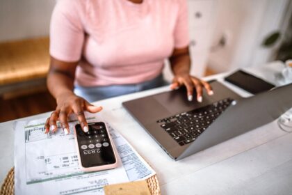 Pessoa usando laptop e smartphone para calcular despesas em planilhas, destacando gestão financeira pessoal.