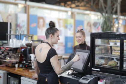 Barista atende cliente em cafeteria moderna, usando tela sensível ao toque; bolos e sanduíches em vitrine ao lado.