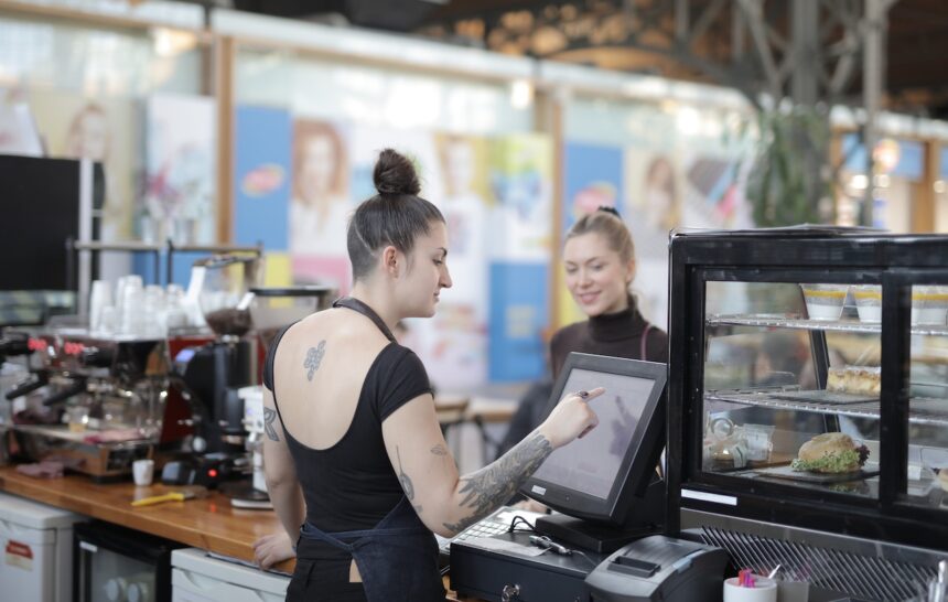 Barista atende cliente em cafeteria moderna, usando tela sensível ao toque; bolos e sanduíches em vitrine ao lado.