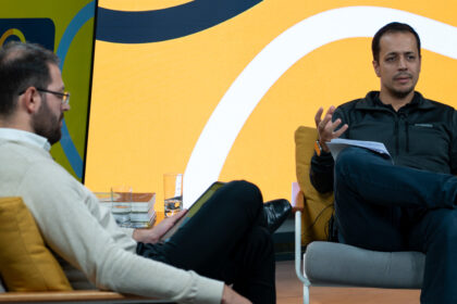 Dois homens em discussão animada, sentados em poltronas modernas, com fundo colorido e livros sobre a mesa.