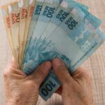 Mãos segurando diversas notas de real, incluindo cédulas de 100 e 50, representando economia e finanças no Brasil.