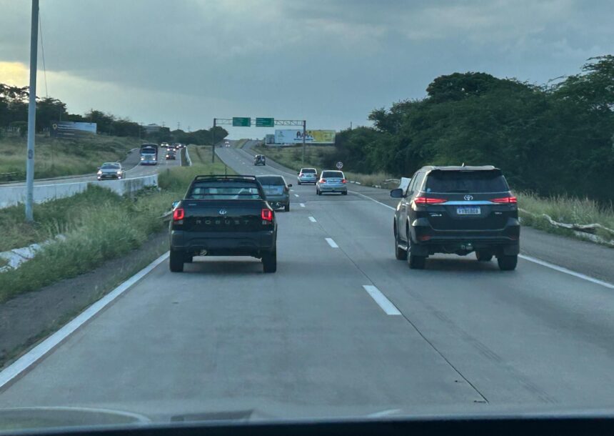 Carros em estrada movimentada, céu nublado ao fundo; vegetação ao lado da via destaca a cena de viagem rodoviária.