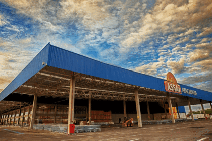 Fachada de um atacadista com cobertura azul, carrinhos de compras e céu nublado ao fundo, destacando o ambiente de compras.