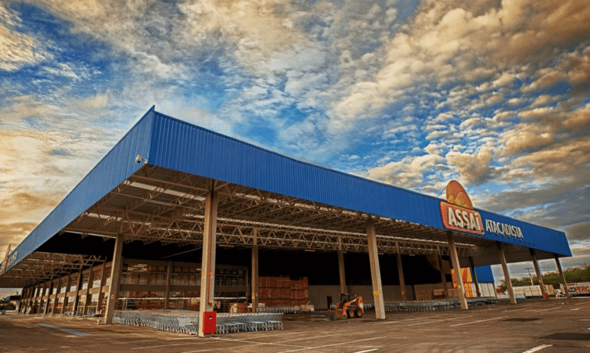 Fachada de um atacadista com cobertura azul, carrinhos de compras e céu nublado ao fundo, destacando o ambiente de compras.