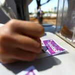 Pessoa preenchendo bilhete de loteria em balcão de quiosque ao ar livre, destacando a aposta na Lotofácil.