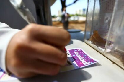 Pessoa preenchendo bilhete de loteria em balcão de quiosque ao ar livre, destacando a aposta na Lotofácil.