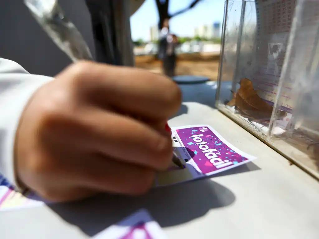 Pessoa preenchendo bilhete de loteria em balcão de quiosque ao ar livre, destacando a aposta na Lotofácil.