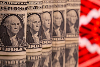Rolos de notas de um dólar lado a lado sobre superfície refletiva, com gráfico vermelho ao fundo, simbolizando economia.
