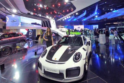 Exposição de carros de luxo com destaque para Porsche 911 GT2RS, em ambiente iluminado, atraindo visitantes.