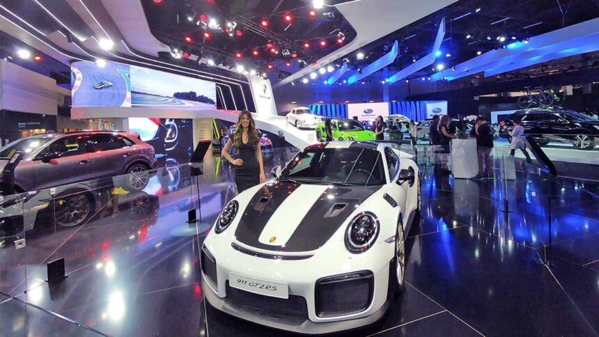 Exposição de carros de luxo com destaque para Porsche 911 GT2RS, em ambiente iluminado, atraindo visitantes.