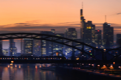 Trem atravessa ponte ao entardecer, com silhuetas de edifícios modernos ao fundo sob um céu alaranjado.