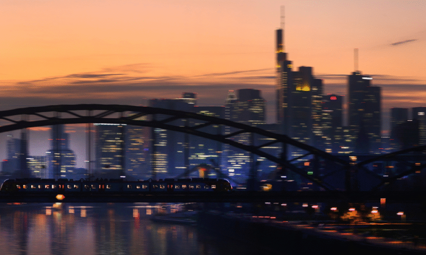Trem atravessa ponte ao entardecer, com silhuetas de edifícios modernos ao fundo sob um céu alaranjado.