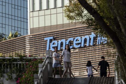 Pessoas passam na frente da sede da Tencent, com logo visível no prédio moderno cercado por vegetação.