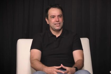 Homem sentado em uma poltrona branca, vestindo camisa preta, em um fundo escuro, parece estar em uma entrevista.
