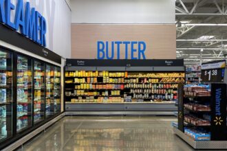 Corredor de supermercado com prateleiras de manteiga e bagels; geladeira de laticínios à esquerda, pães ao lado.