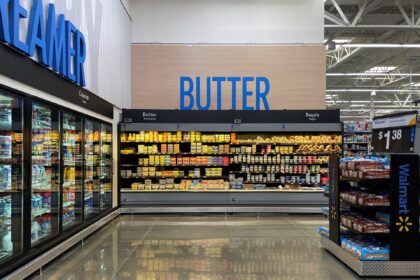 Corredor de supermercado com prateleiras de manteiga e bagels; geladeira de laticínios à esquerda, pães ao lado.