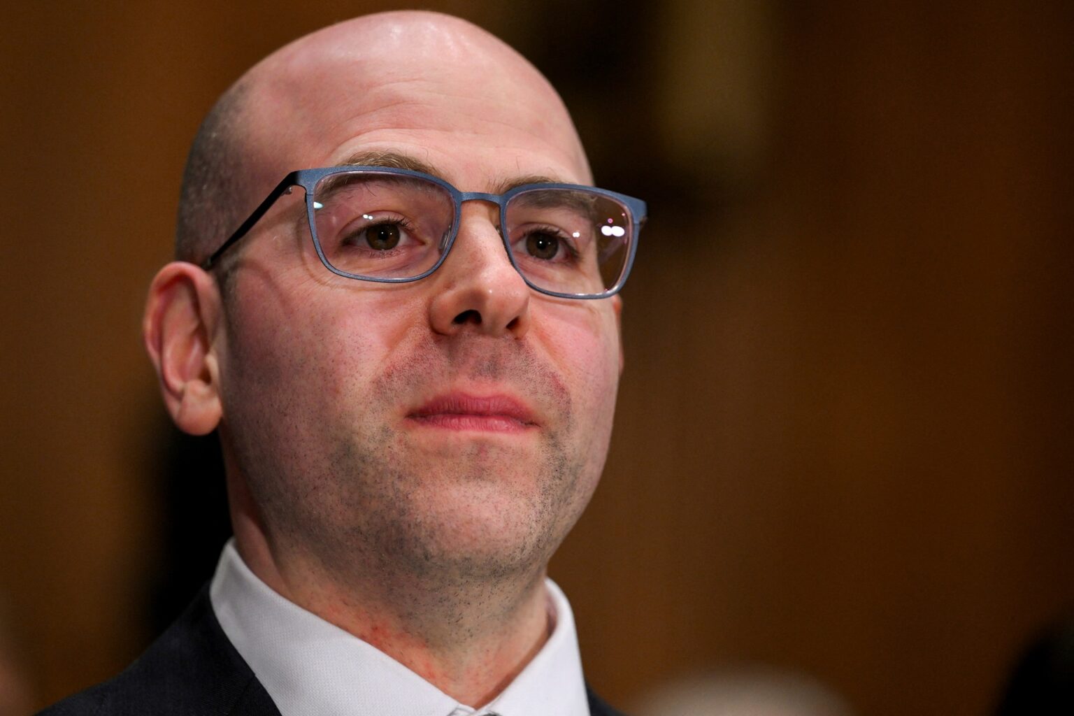 Homem com óculos e terno observa atentamente. Ambiente formal, fundo desfocado. Expressão séria, foco nos detalhes faciais.