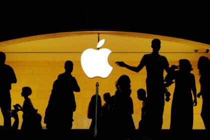 Silhuetas de pessoas em frente a uma loja da Apple, iluminada pelo logotipo brilhante da marca ao fundo.