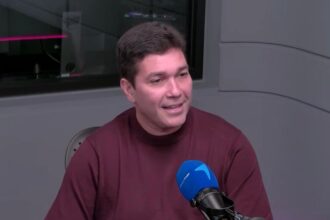 Homem sorrindo em estúdio de gravação, vestindo suéter marrom, frente a um microfone azul, com fundo cinza moderno.