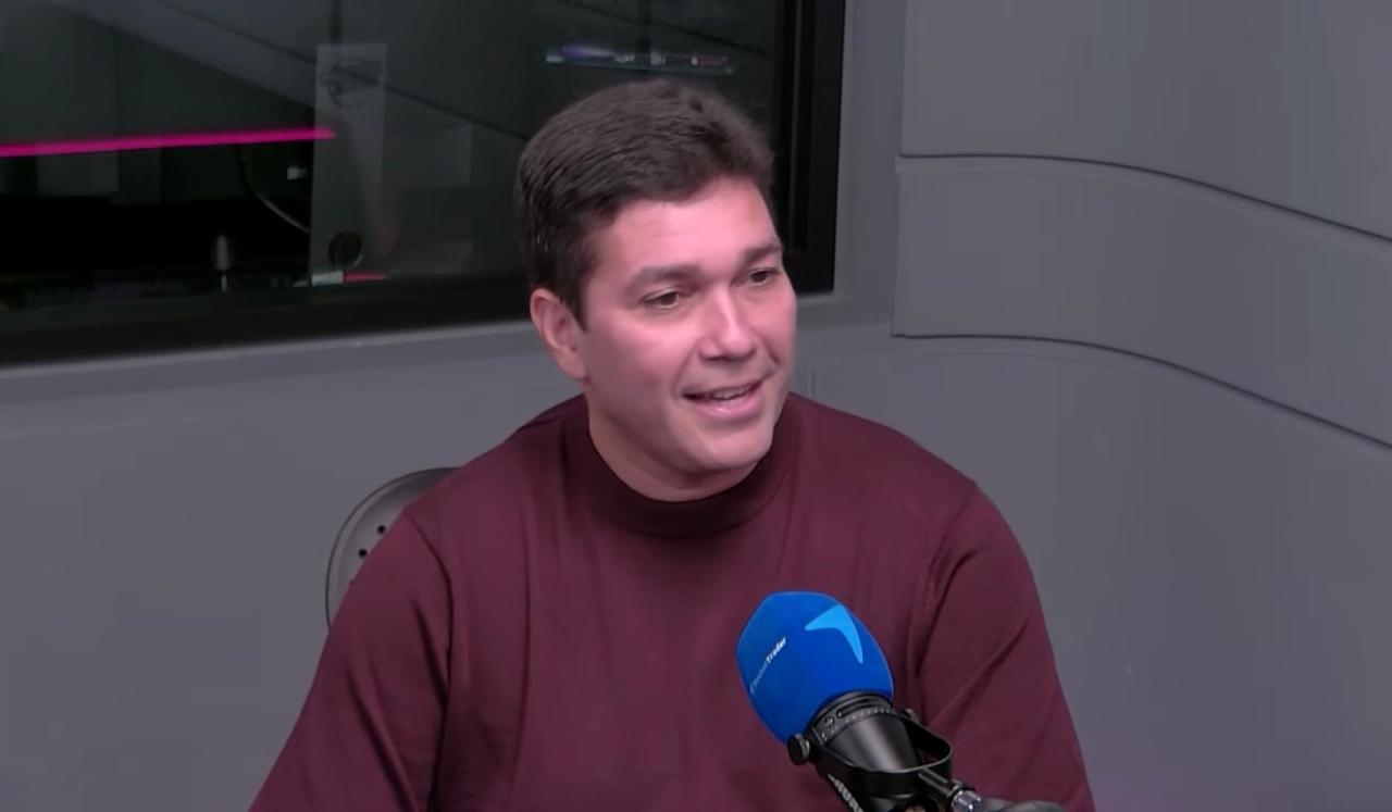 Homem sorrindo em estúdio de gravação, vestindo suéter marrom, frente a um microfone azul, com fundo cinza moderno.