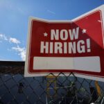 Placa de Now Hiring pendurada em uma cerca sob céu azul, simbolizando oportunidades de emprego disponíveis.