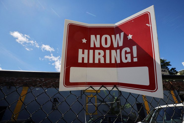 Placa de Now Hiring pendurada em uma cerca sob céu azul, simbolizando oportunidades de emprego disponíveis.