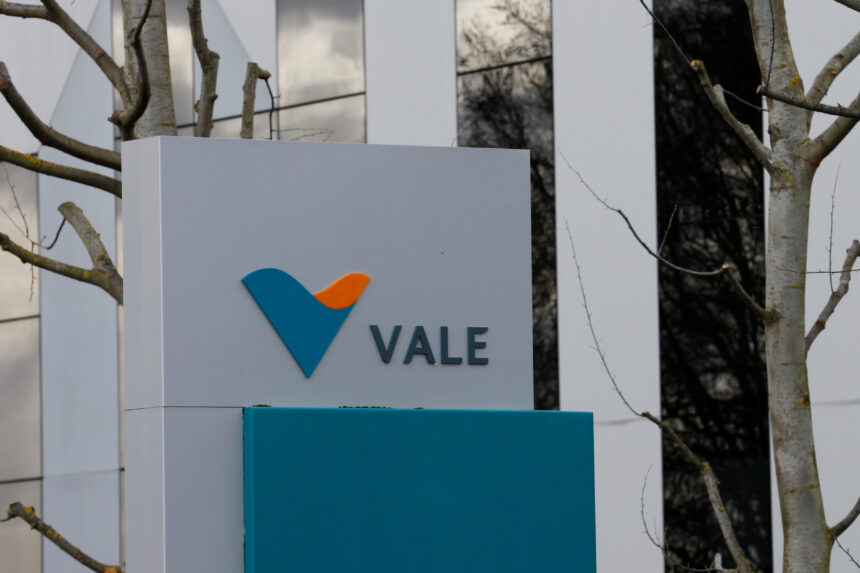 Placa com o logotipo da empresa Vale em um ambiente externo, com árvores ao fundo e prédio moderno refletido.