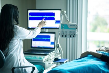 Profissional de saúde monitora paciente deitado, analisando dados em telas de computador em ambiente hospitalar.