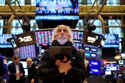 Homem observa atentamente as telas de cotações na Bolsa de Valores, segurando um tablet enquanto analisa dados financeiros.