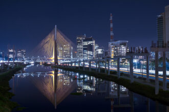 Ponte estaiada iluminada à noite em São Paulo, refletida no rio, com prédios modernos ao fundo. Trânsito intenso nas margens.