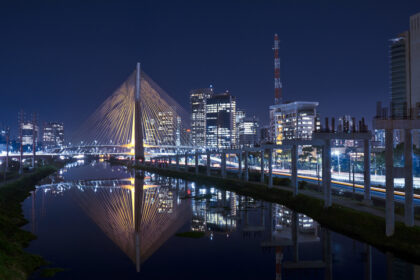 Ponte estaiada iluminada à noite em São Paulo, refletida no rio, com prédios modernos ao fundo. Trânsito intenso nas margens.