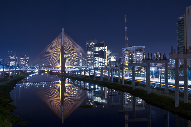 Ponte estaiada iluminada à noite em São Paulo, refletida no rio, com prédios modernos ao fundo. Trânsito intenso nas margens.