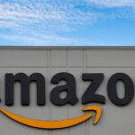 Logo tridimensional da Amazon em fachada de edifício cinza, com céu azul e nuvens ao fundo.