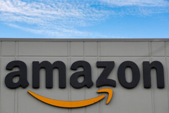 Logo tridimensional da Amazon em fachada de edifício cinza, com céu azul e nuvens ao fundo.