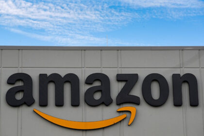 Logo tridimensional da Amazon em fachada de edifício cinza, com céu azul e nuvens ao fundo.