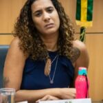 Uma mulher com expressão séria está sentada em uma sala, ao lado de uma garrafa rosa e um copo d'água, com uma bandeira ao fundo.