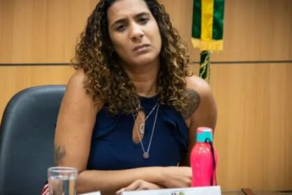 Uma mulher com expressão séria está sentada em uma sala, ao lado de uma garrafa rosa e um copo d'água, com uma bandeira ao fundo.