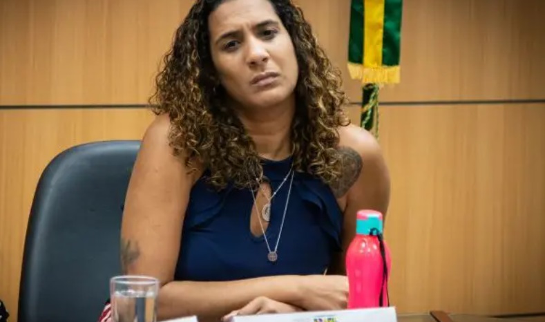 Uma mulher com expressão séria está sentada em uma sala, ao lado de uma garrafa rosa e um copo d'água, com uma bandeira ao fundo.