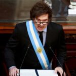 Político argentino faz discurso enérgico, usando faixa presidencial azul e branca, em ambiente governamental com microfones.